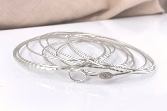 A stack of thin sterling silver bangle bracelets with various textures, including a twist design, on a white background.