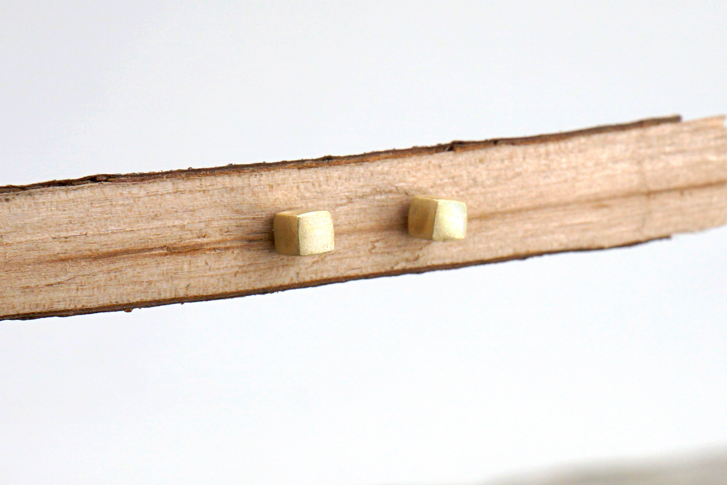 Two gold square stud earrings on a wooden stick against a white background